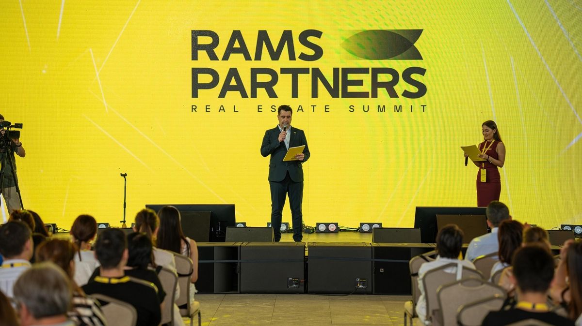 Джем Джиритджи, змдиректора RAMS Qazaqstan, человек с микрофоном на сцене с желтым фоном перед залом со зрителями