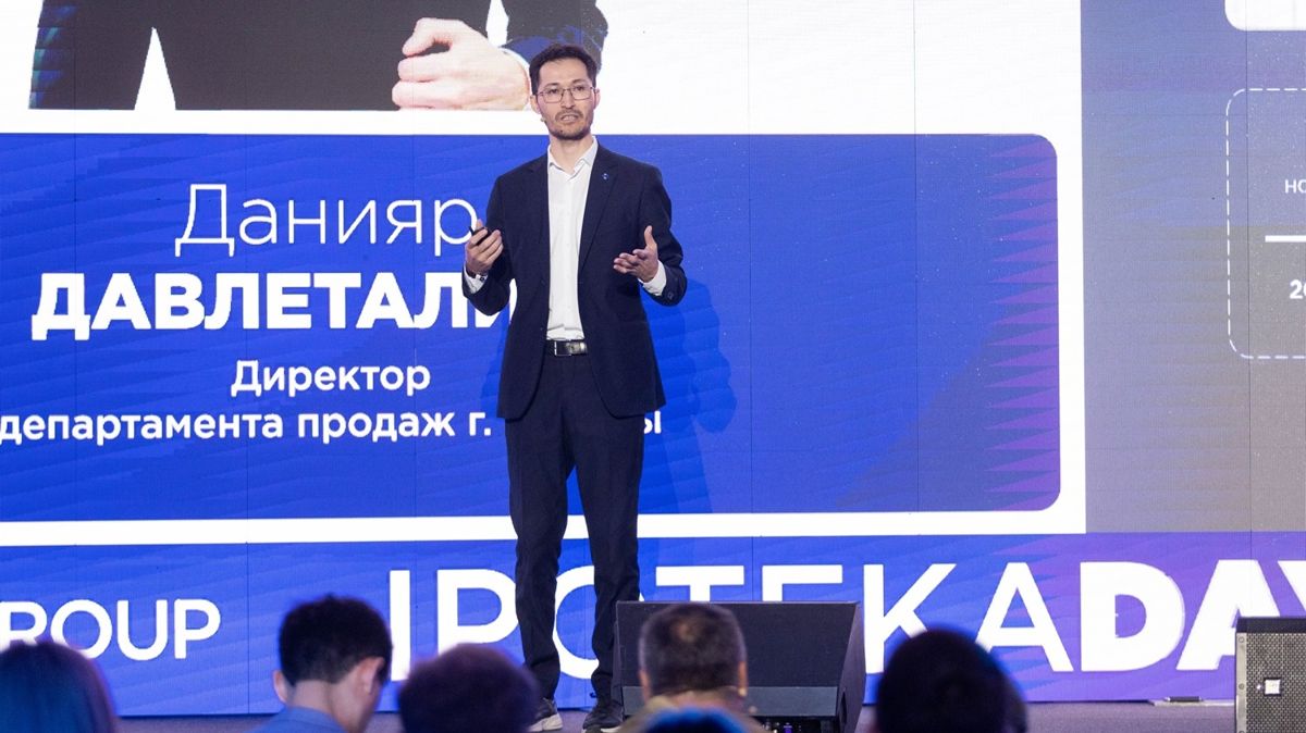 Данияр Давлеталиев, мужчина стоит на сцене на фоне экрана, где написано его имя