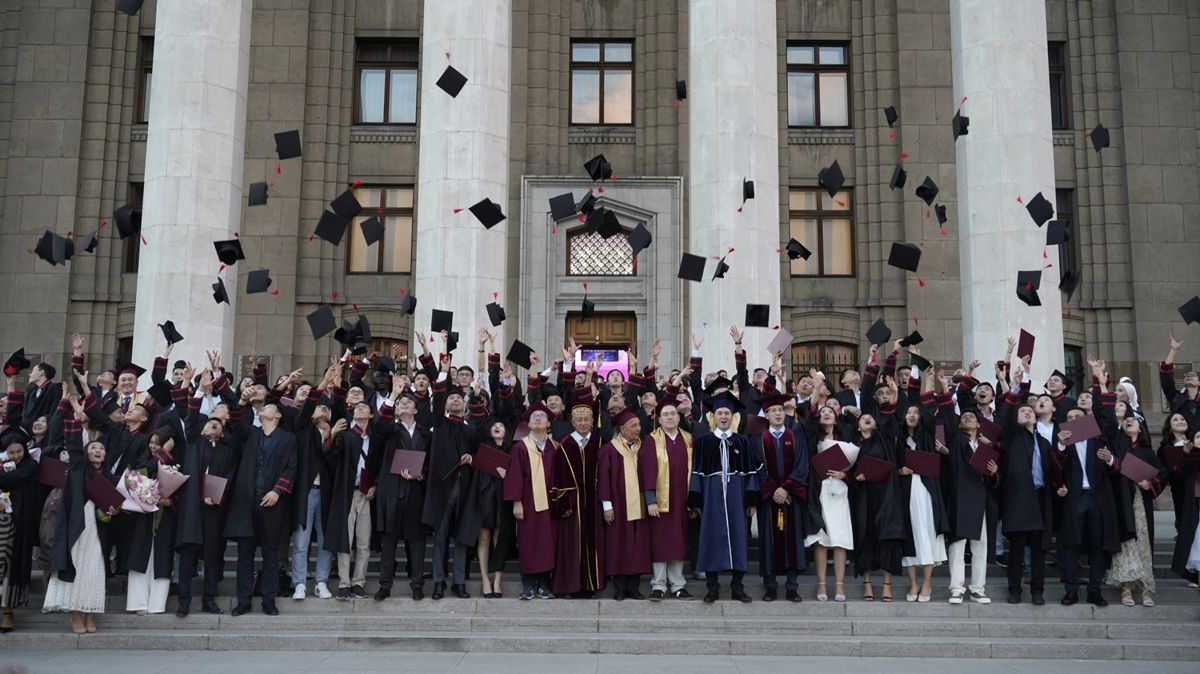 Выпускники КБТУ на ступенях здания с колоннами бросают вверх академические шапочки
