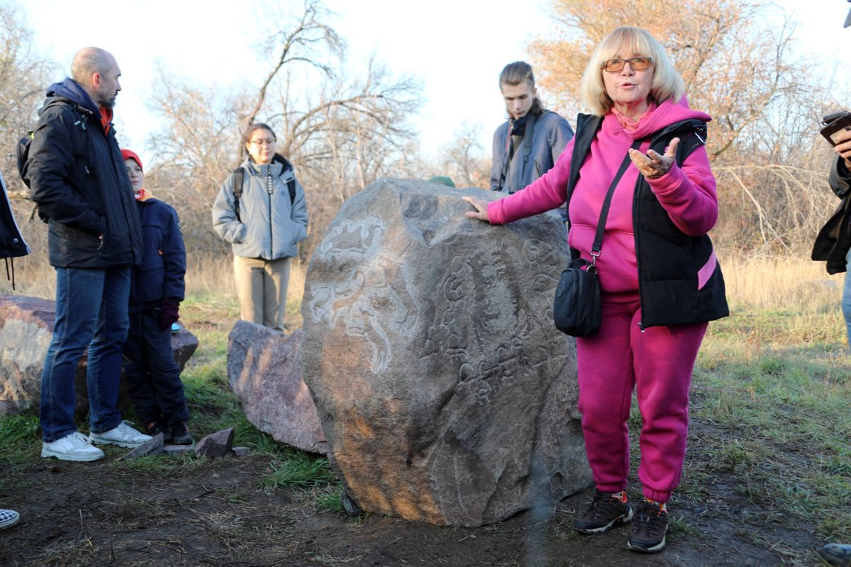 Женщина в розовой куртке показывает на камень с петроглифами