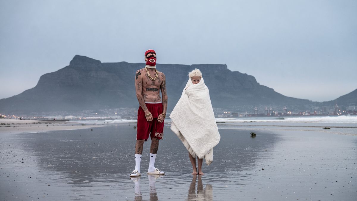 Die Antwoord создали абсолютно новый стиль Мужчина с голым торсом в красных шортах и в красной маске на лице и женщина в белом палантине