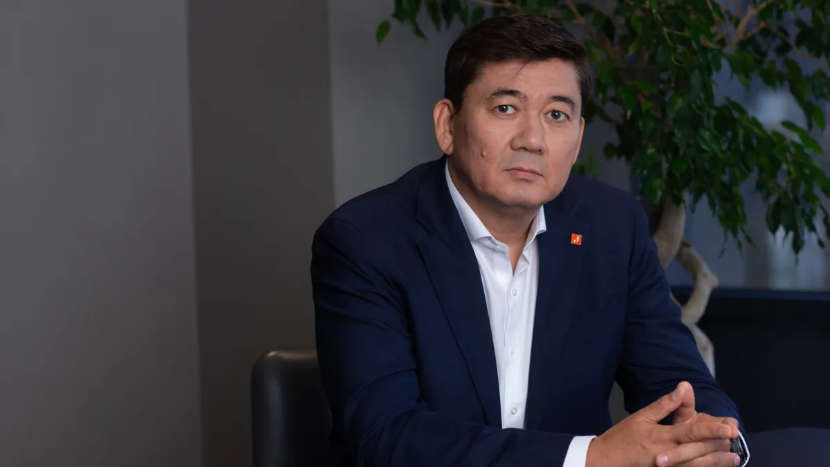 Куат Кожахметов в темно-синем пиджаке и белой рубашке без галстука сидит за столом