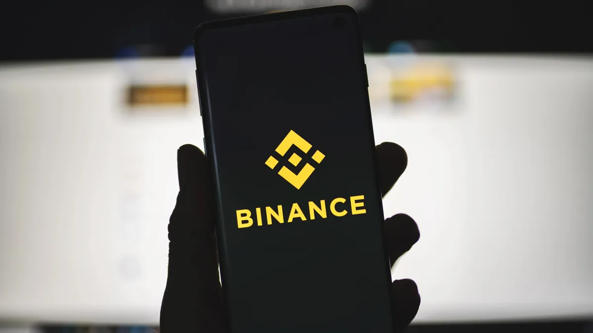 Человек держит смартфон с логотипом Binance с размытым веб-сайтом в качестве фона. Обмен криптовалютами, торговая платформа